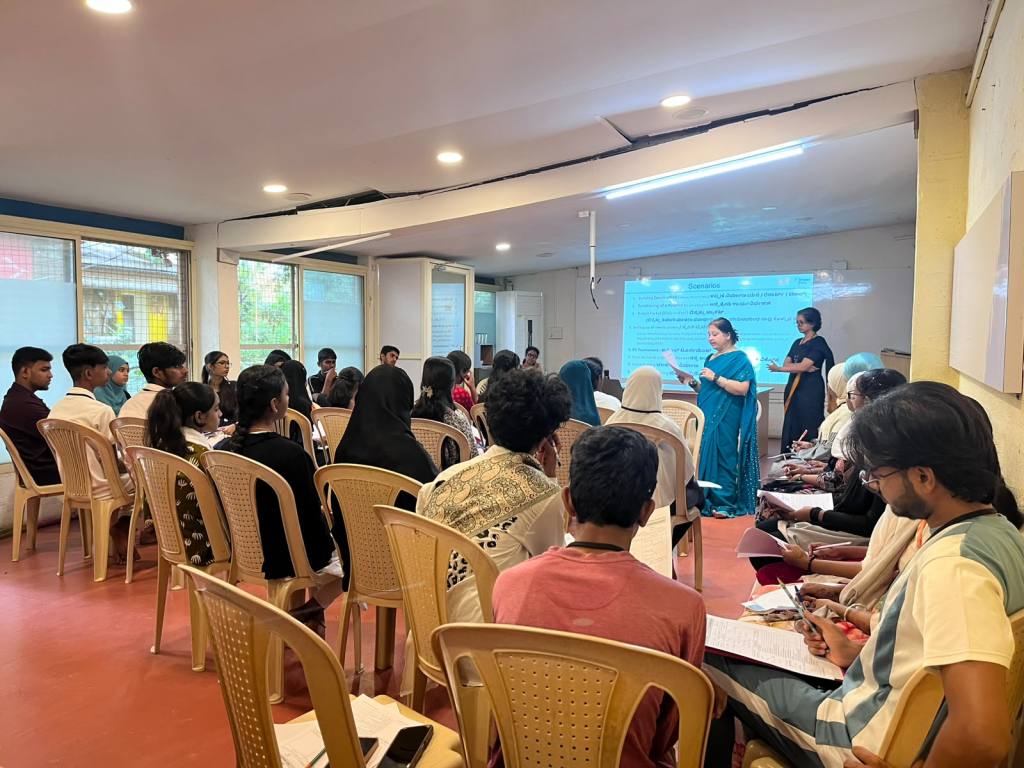 A group of students attending a workshop in a classroom setting, with a female instructor in a teal sari speaking while another instructor supports her, and attendees taking notes.